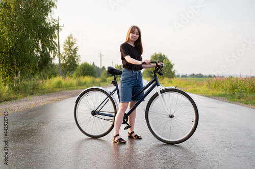 Young woman standing alone on road with bicycle looking at camera at countryside. Summer activity, healthy lifestyle, workout, sport, fitness. Person in nature. Riding bicycle. Having fun outdoors.