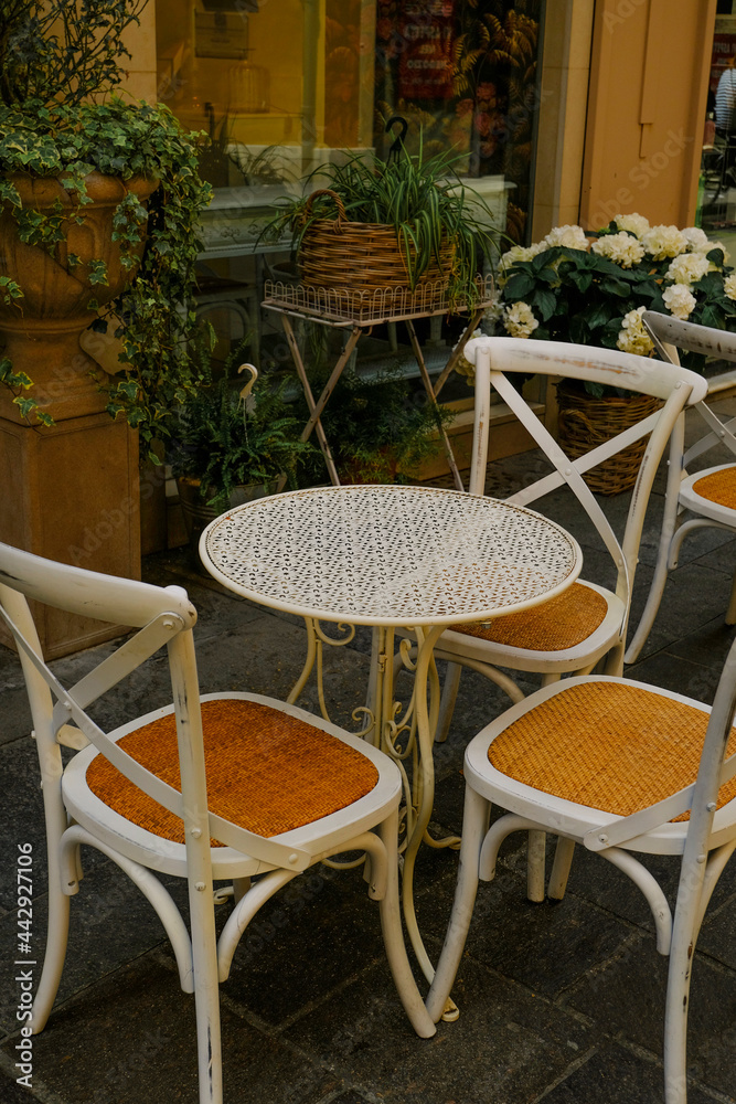White wooden table with chairs across plants in pots in restaurant terrace. Cafe exterior. City life
White wooden table with chairs across plants in pots in restaurant terrace. Cafe exterior. City life