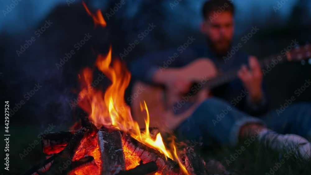 Beautiful campfire at night. Red and orange flame burning on blur background of a man playing the guitar in the evening. Leisure time outdoors. Beautiful campfire at night. Red and orange flame burning on blur background of a man playing the guitar in the evening. Leisure time outdoors.