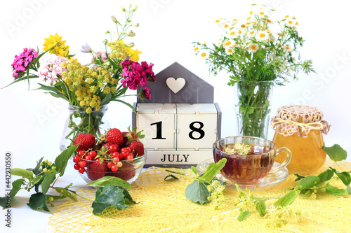 Calendar for July 18 : the name of the month of July in English, cubes with the number 18, bouquets of wild flowers, jam, fruit, a cup of tea on a yellow openwork napkin © MARYIA
