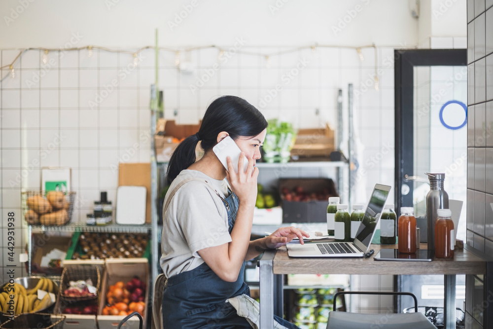 Kvinnlig entreprenör pratar i telefon och arbetar på laptop sittande i butik Kvinnlig entreprenör pratar i telefon och arbetar på laptop sittande i butik