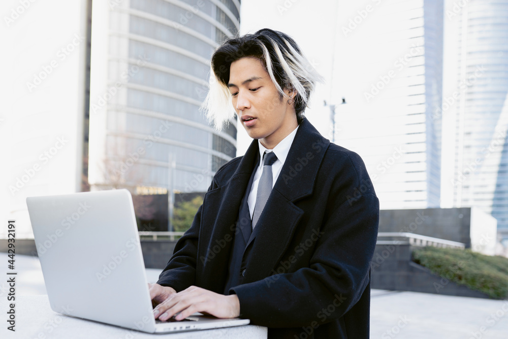 Stylish Asian entrepreneur typing on laptop against urban skyscrapers Stylish Asian entrepreneur typing on laptop against urban skyscrapers