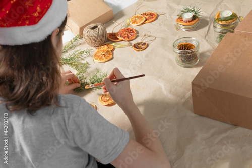 A back view of teen girl  in Santa het making decoration  and candles for Christmas gifts,  kraft beige paper box with dry slices of gold orange, grapefruits, green pine branch. Handmade  concepts. photo