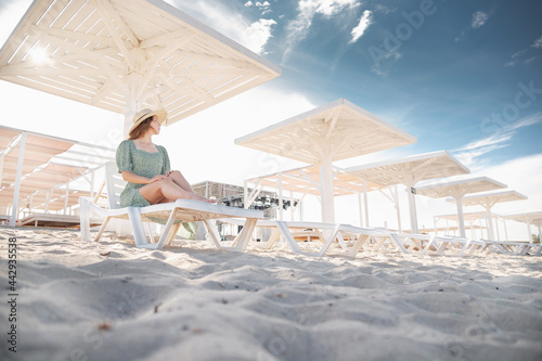 A blonde in a green dress sits on a sunbed and looks at the seascape under protective sunny wooden umbrellas. Young caucasian attractive woman by the sea on the beach