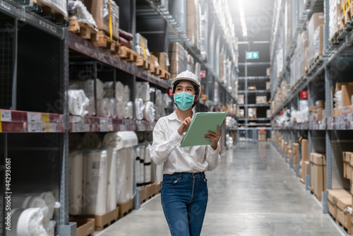 Engineer woman worker wearing mask and safety helmet doing stocktaking of product management in cardboard box on shelves in warehouse. Factory physical inventory count.