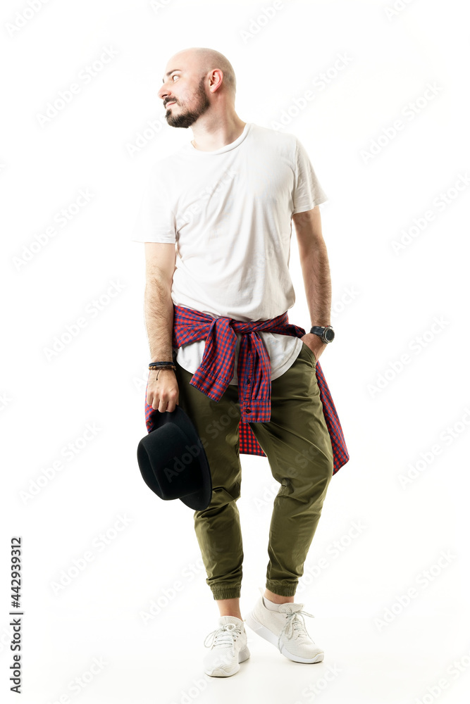 Stylish shaved bearded hipster holding hat looking away with plaid shirt tied at waist. Full body portrait isolated on white background Stylish shaved bearded hipster holding hat looking away with plaid shirt tied at waist. Full body portrait isolated on white background
