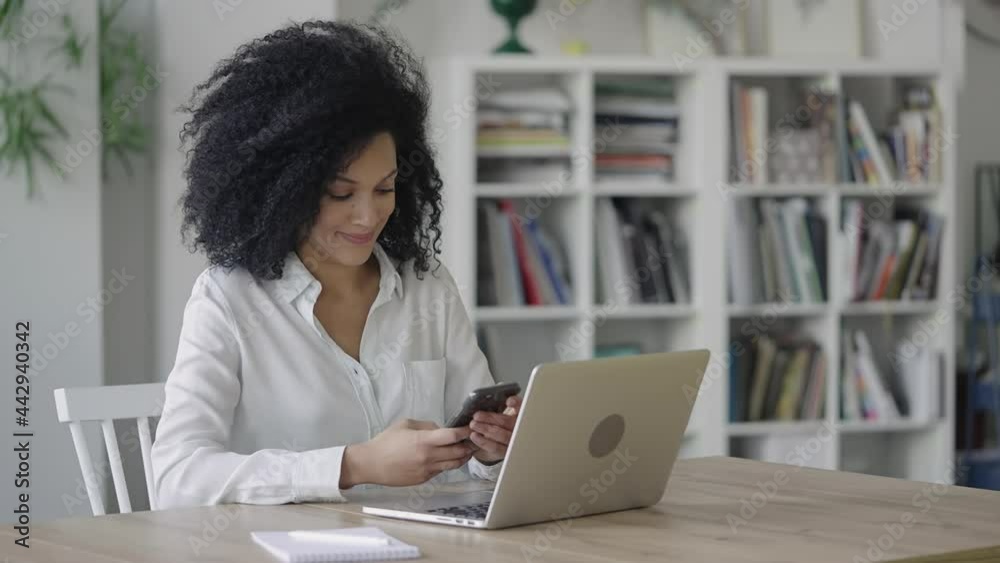 Portrait of a young African American woman texting on a smartphone. A brunette with curly hair in a white blouse sits at a table in a light home office. Close up. Slow motion ready 59.97fps. Portrait of a young African American woman texting on a smartphone. A brunette with curly hair in a white blouse sits at a table in a light home office. Close up. Slow motion ready 59.97fps.