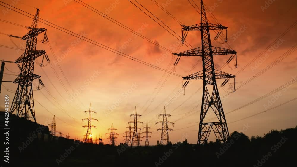 Supports of high-voltage power lines against the backdrop of red sunset. Sun setting and power line pylon silhouettes in epic sunset sky.