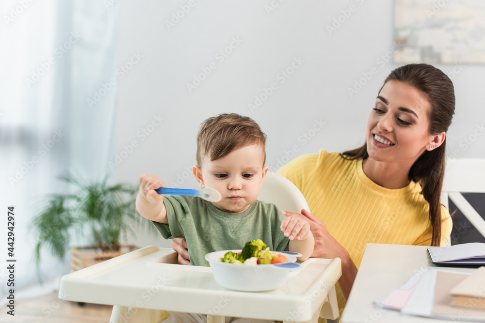 Smiling mother looking at son with spoon near vegetables in bowl on high chair