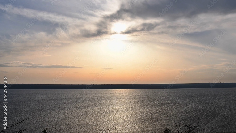 White sun shines in high sky with light spindrift clouds over tranquil water surface. Sunshine over shore landscape with blurry sunlight reflection