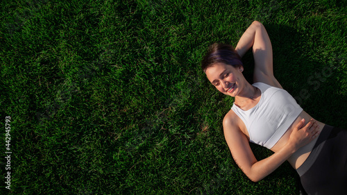 A beautiful, cheerful, cheerful woman in a white T-shirt is lying on the green lawn in the park and smiling. Happy woman relaxing on the grass during sunny summer day. Top view. large format banner