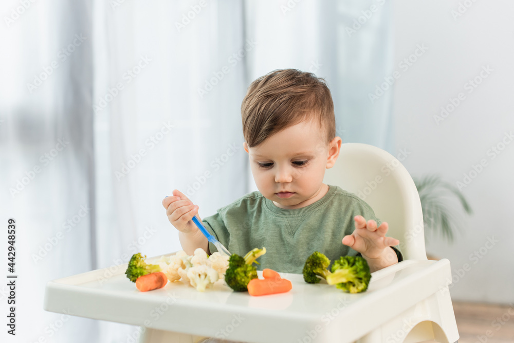 Kid with spoon looking at vegetables on high chair at home Kid with spoon looking at vegetables on high chair at home