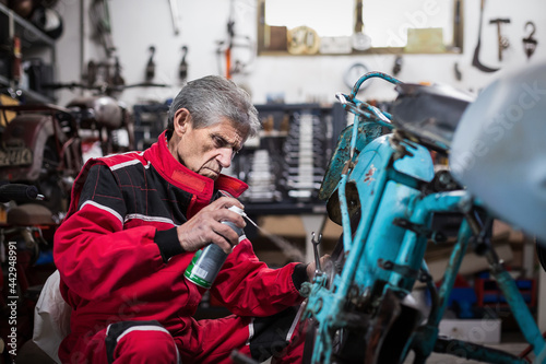 Elderly mechanic spraying oil on motorcycle detail in workshop photo