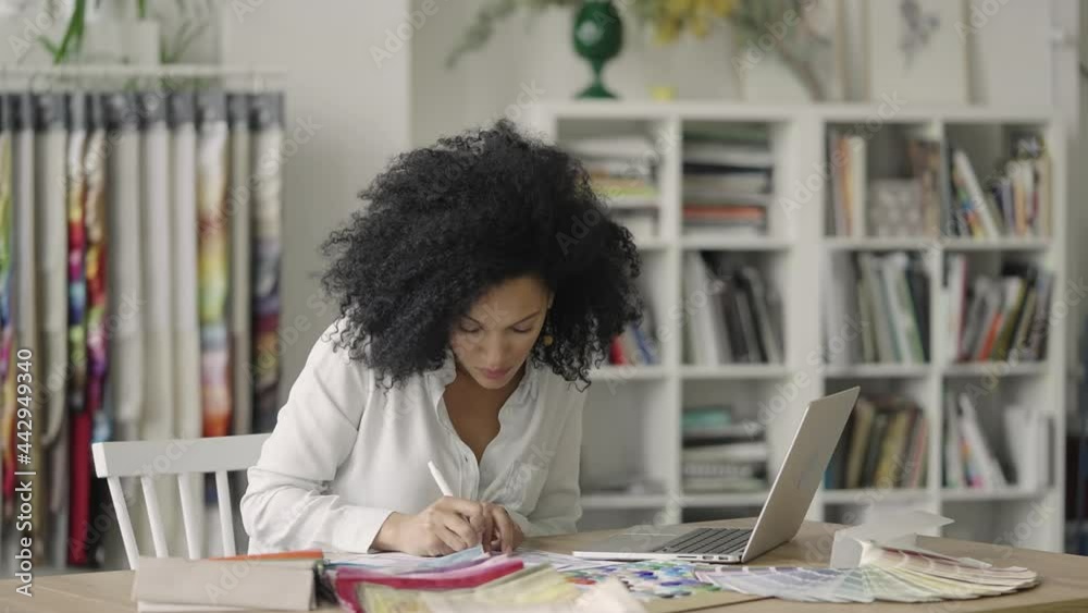 Portrait of young African American woman looking through color palette and samples of fabrics for design project. Female designer sits at table in office. Close up. Slow motion ready 59.97fps. Portrait of young African American woman looking through color palette and samples of fabrics for design project. Female designer sits at table in office. Close up. Slow motion ready 59.97fps.