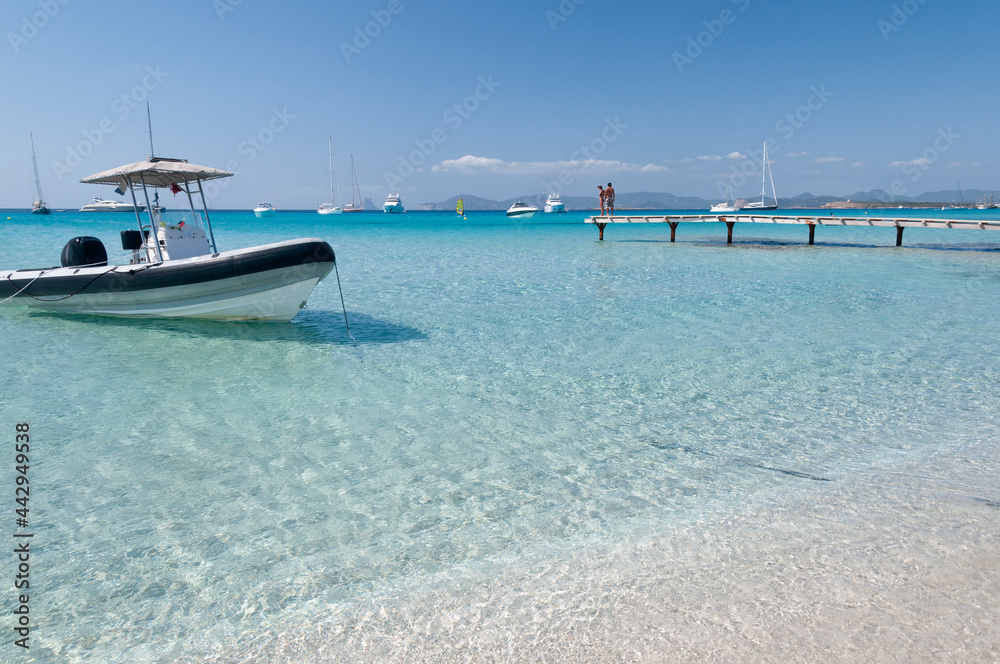 Fototapeta premium A zodiac on the beach of Illetas, the island of Formentera in Spain. In the background the blue sky and a wooden pier with tourists. 