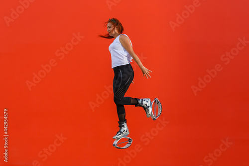 Latin woman doing a jump while flexing one leg backwards wearing Kangoo Jumps boots. Red haired woman doing an exercise routine