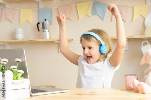Smiling little happy schoolgirl raises her hands to celebrate the victory. little fan is celebrating a victory.Laptop online lesson video call listen to good news good grades for the semester 