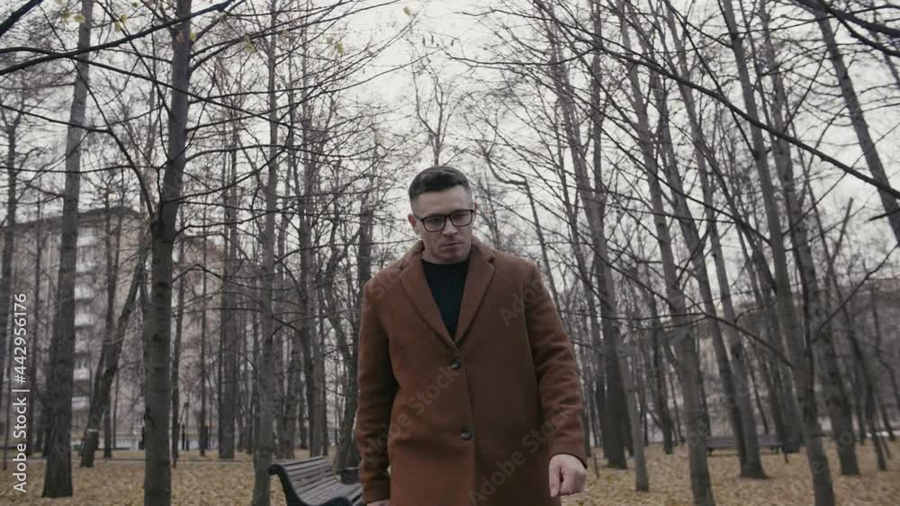 Handsome young man in warm coat walking on street during autumn weather. Frustrated young guy walks through park in city center. Guy with glasses enjoying fresh air outdoors Handsome young man in warm coat walking on street during autumn weather. Frustrated young guy walks through park in city center. Guy with glasses enjoying fresh air outdoors