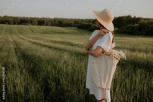 A girl in a hat and a white dress stands in a green field of wheat on her hand hangs a sumka with a bouquet of daisies photo