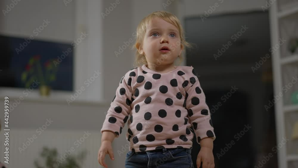 Medium shot portrait of charming Caucasian baby girl standing in living room looking away. Pretty blond child at home in the evening. Infantry concept Medium shot portrait of charming Caucasian baby girl standing in living room looking away. Pretty blond child at home in the evening. Infantry concept