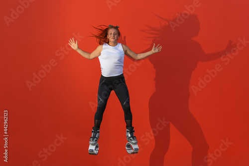 Latin, redhead woman in sportswear jumping with open arms wearing Kangoo Jumps boots on orange background. Active movement, action, fitness and wellness concept