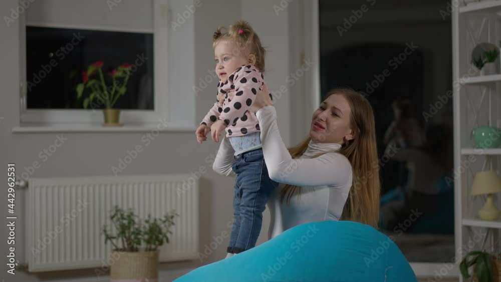 Side view of young millennial mother raising baby girl smelling diaper. Beautiful slim Caucasian woman taking care of daughter at home indoors