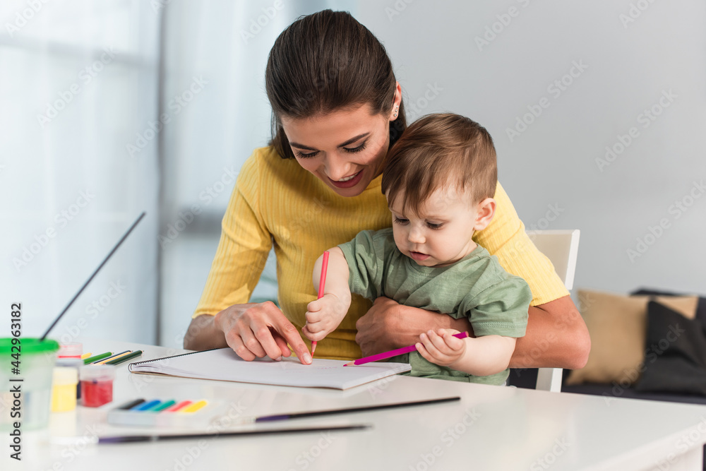 Young woman smiling while pointing at paper near son with pencils at home