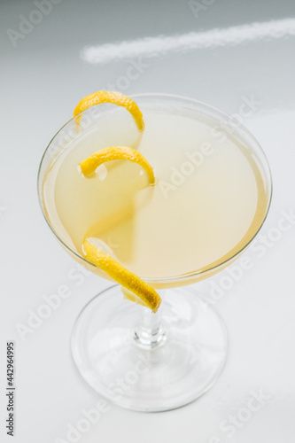yellow cocktail in coupe glass with long curly lemon twist against white background