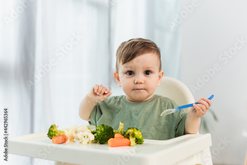 Toddler boy holding spoon and looking at camera near vegetables on high chair