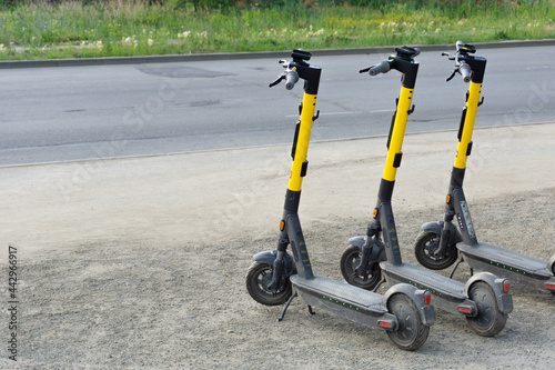 Electric scooters are parked by the road. Yellow and black scooters stand near the park. Eco-friendly transport.