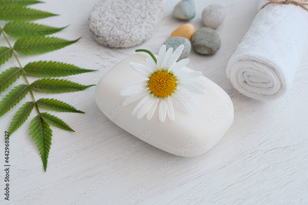 White soap with a chamomile flower on top in a blue soap dish next to green leaves, with a towel and stones of the sea. Spa concept, treatments, body hygiene, natural cosmetics White soap with a chamomile flower on top in a blue soap dish next to green leaves, with a towel and stones of the sea. Spa concept, treatments, body hygiene, natural cosmetics