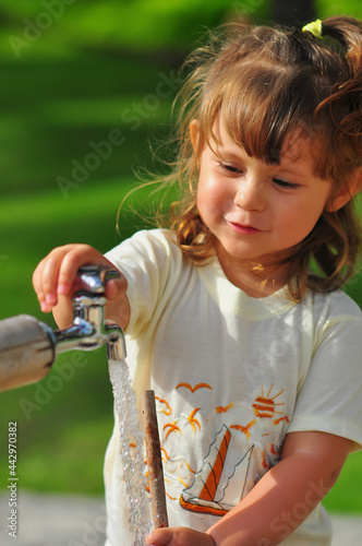 Portrait of a little caucasian girl opening a water tap in a park near the sea beach. Outdoors on the background of a green lawn. Summer sunny day. Healthy lifestyle concept.