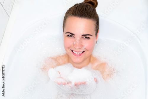 Beauty portrait of beautiful happy positive girl, young cheerful woman is relaxing, lying in bathroom, taking a bath with bubbles, smiling and looking at camera, washing her body with foam in bathtub
