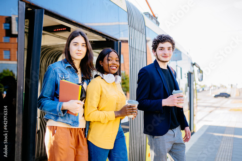 Mixed raced group of friends millennial students on tram stop. Lifestyle photo of young women and man come out while opening door to tram. Portrait of female student In denim jacket with books smiling photo