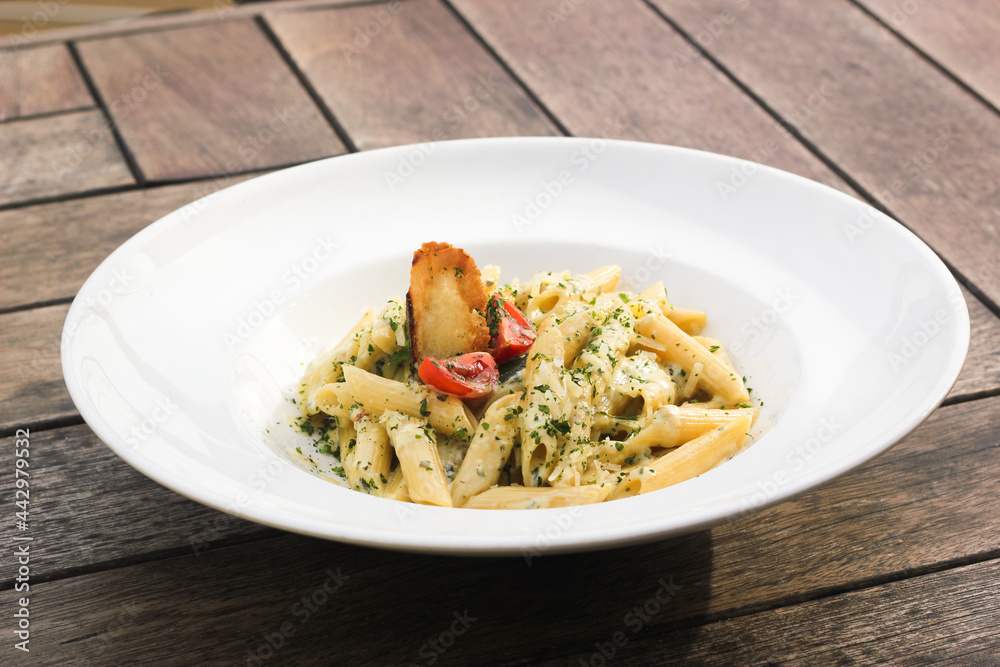Four cheese penne pasta with basil and tomato in white plate on wooden table. Italian pasta. Pesto souce.