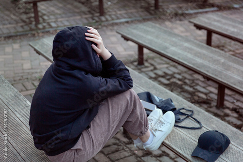 A teenage girl in a black hoodie with a hood on her head is sitting on a bench, covering her face with her hands, back view. Life and problems of teenagers, loneliness concept