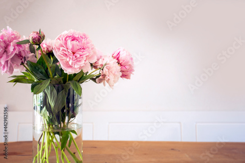 A bunch of pastel pink peony with green leaves in glass vase on the wooden table near white wall texture, cozy bright decoration for stylish house with copy space