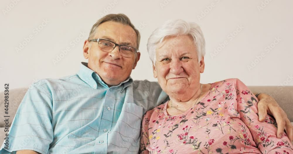 A long married couple, relaxing together at home on a sofa, smiling pensioners, a couple in love, a grandfather embracing a grandmother, looking at the camera, happy, cheerful A long married couple, relaxing together at home on a sofa, smiling pensioners, a couple in love, a grandfather embracing a grandmother, looking at the camera, happy, cheerful