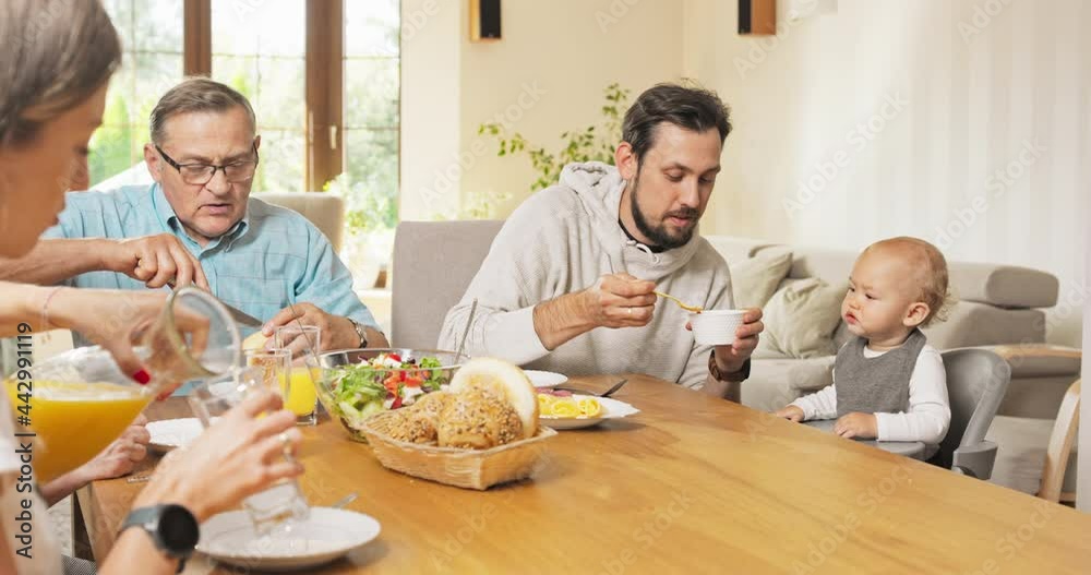 Family breakfast at home at table A man feeds his little son sitting in a chair, the child eats with appetite, a woman next to him pours orange juice into a glass. The grandfather is cutting bread Family breakfast at home at table A man feeds his little son sitting in a chair, the child eats with appetite, a woman next to him pours orange juice into a glass. The grandfather is cutting bread