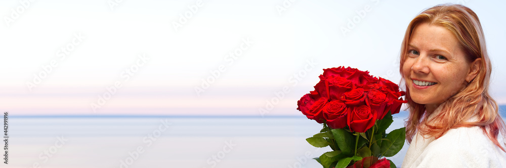 happy beautiful woman 40s in a white robe with a bouquet of red roses looks from the hotel balcony at the adreatic sea. romantic vacation, celebrating birthday. copy space. Spa hotel resort. luxury