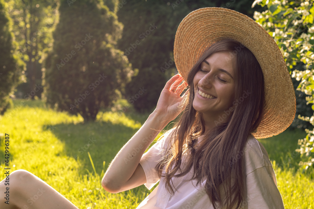 Happy and smiling young woman wearing sun hat relaxes in nature enjoying weekend on a sunny day. Woman happy and joyful, wellbeing and relaxation.
