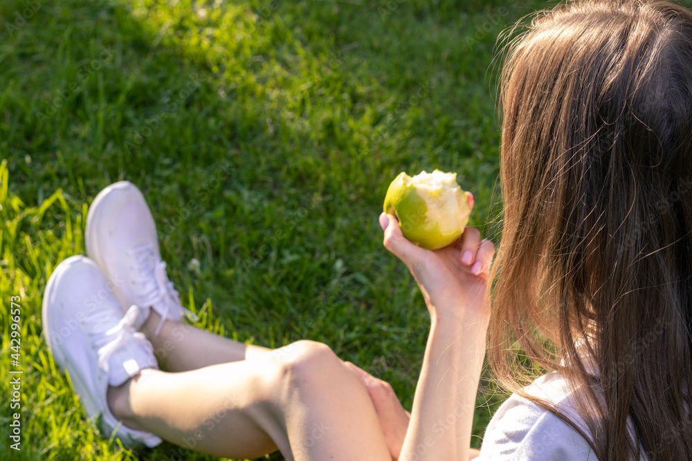 Young woman relax in nature on a sunny day and eats green apple; woman sits on green grass and holds bitten apple in her hands; back photo. Young woman relax in nature on a sunny day and eats green apple; woman sits on green grass and holds bitten apple in her hands; back photo.