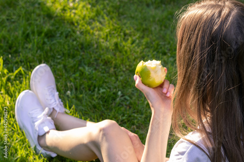 Young woman relax in nature on a sunny day and eats green apple; woman sits on green grass and holds bitten apple in her hands; back photo.