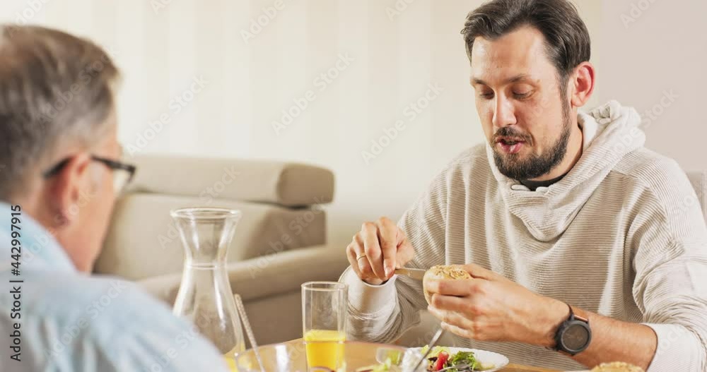 A handsome young brunet sits with his father at the table, they eat breakfast together, the man cuts a roll, tells his father about work, home, children, laughs, his father gives him butter A handsome young brunet sits with his father at the table, they eat breakfast together, the man cuts a roll, tells his father about work, home, children, laughs, his father gives him butter