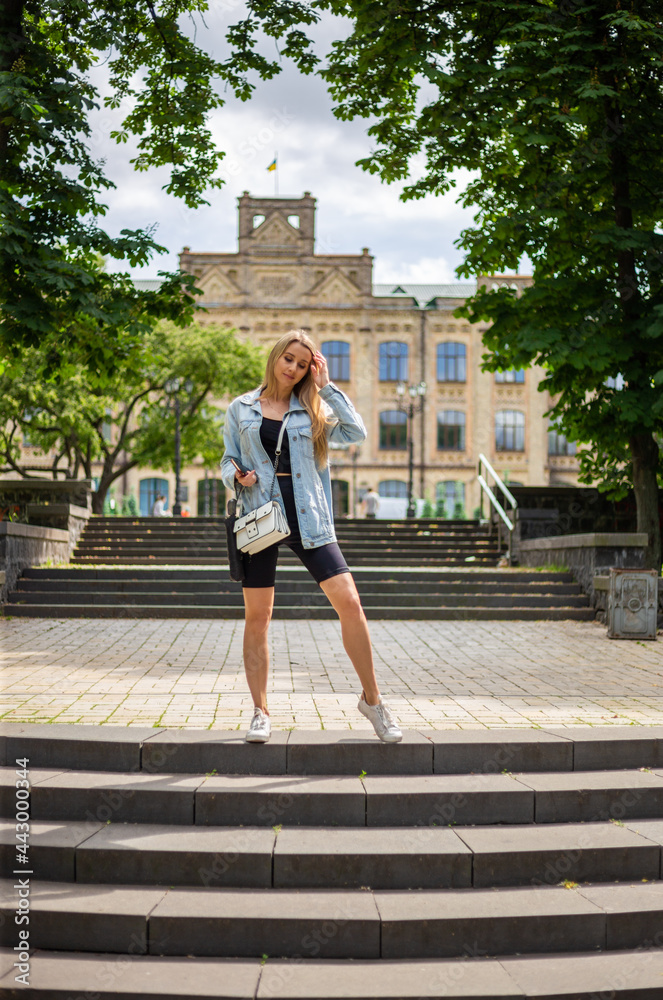 Young beautiful blonde woman in a denim jacket and tight sports shorts stands on the stairs