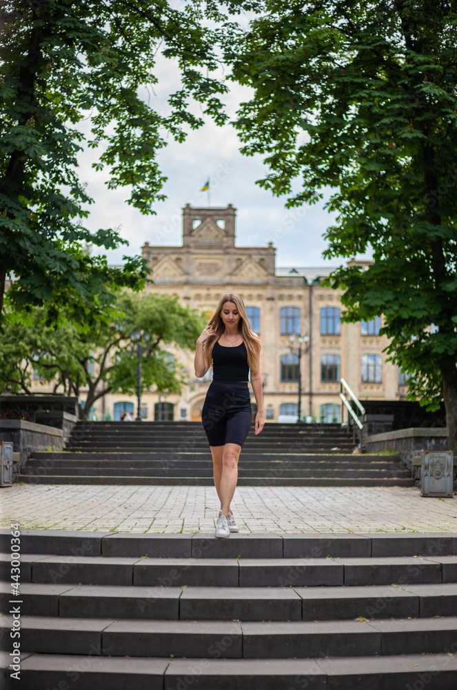Young beautiful blonde woman with athletic build in a black t-shirt and in black tight sports shorts stands on the stairs a Young beautiful blonde woman with athletic build in a black t-shirt and in black tight sports shorts stands on the stairs a