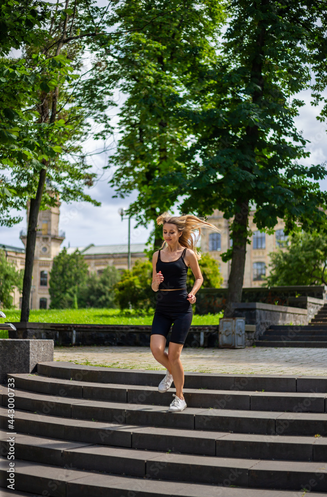 A young blonde woman with a sporty physique in a black T-shirt and in black tight sports shorts runs up the stairs