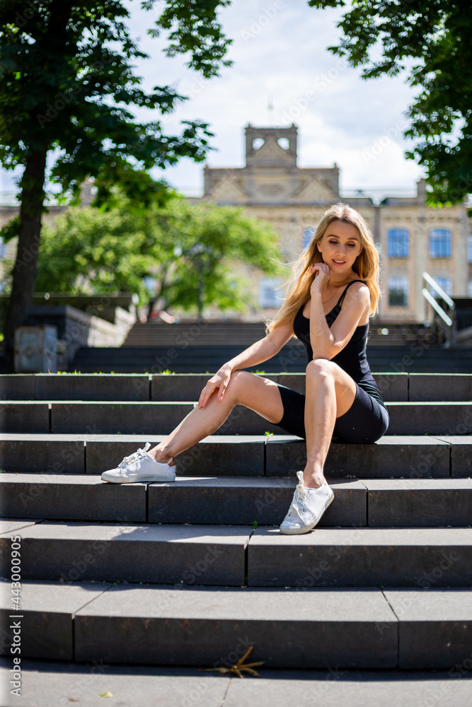 Young blonde sportive woman in a black tank top and in black tight sports shorts sits on the stairs Young blonde sportive woman in a black tank top and in black tight sports shorts sits on the stairs