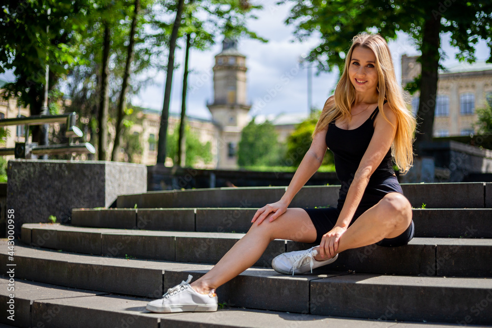 Young blonde sportive woman in a black tank top and in black tight sports shorts sits on the stairs