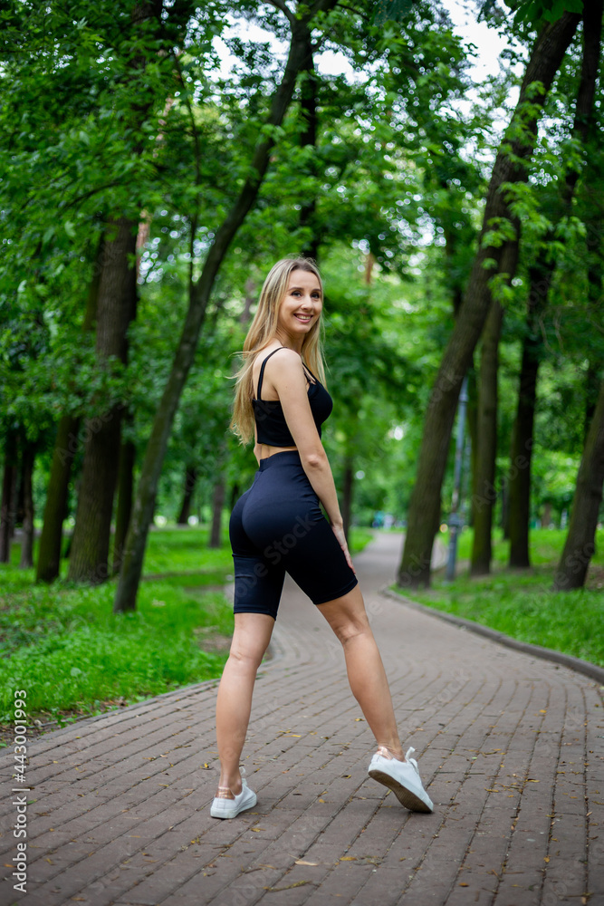 Young beautiful sporty blonde woman in a black T-shirt and in black tight sports shorts in good shape posing in front of green trees Young beautiful sporty blonde woman in a black T-shirt and in black tight sports shorts in good shape posing in front of green trees